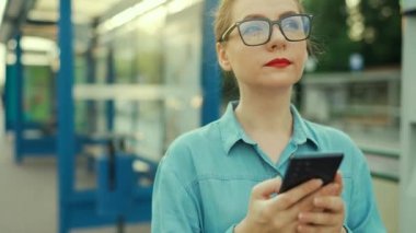 Woman stands at a public transport stop, using smartphone and waiting for the tram. City, urban, transportation. Slow motion