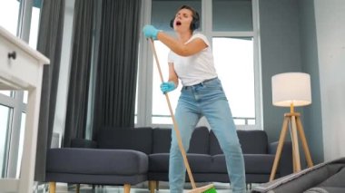 Beyaz kulaklıklı kadın evi temizliyor ve konserdeki bir yıldız gibi süpürgeyle şarkı söyleyerek eğleniyor. Yavaş çekim