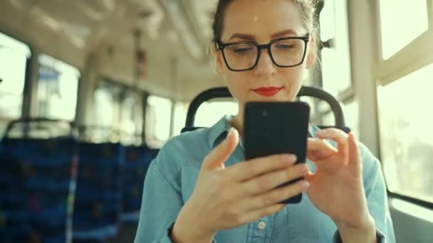 公共交通機関だ スマートフォンのチャットや友人とのテキストメッセージを使用してトラムのメガネの女性 — ストック動画
