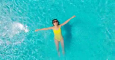 Sarı mayo giymiş bir kadının havuzda sırt üstü yatışı. Rahatlatıcı konsept, yaz yaşam tarzı. Kamera suyun üzerinde yükseliyor.