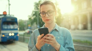 Kadın bir toplu taşıma durağında duruyor, akıllı telefon kullanıyor ve tramvayı bekliyor. Şehir, şehir, ulaşım. 