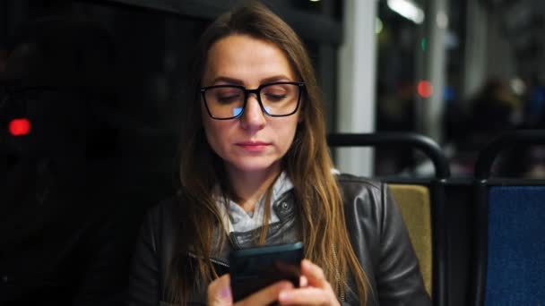 公共交通工具在夜间 坐在电车里戴眼镜的女人用智能手机和朋友聊天和发短信 — 图库视频影像