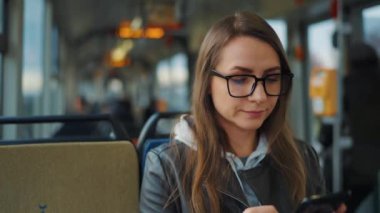 Public transport. Woman in glasses in tram using smartphone chatting and texting with friends, slow motion. City, urban, transportation.