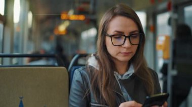 Public transport. Woman in glasses in tram using smartphone chatting and texting with friends, slow motion. City, urban, transportation.