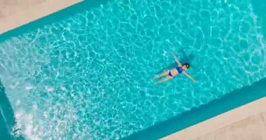 Top down view of a woman in a blue swimsuit lying on her back in the pool, slow motion. Relaxing concept, summer lifestyle. Camera rises and rotates clockwise