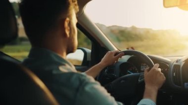 Adam yol boyunca araba sürüyor, batan güneş ön camda parlıyor. Sürücülerin omzunun arkasından bak.