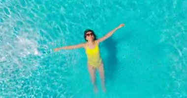 Top down view of a woman in an yellow swimsuit lying on her back in the pool and camera rising fast. Relaxing concept, summer lifestyle