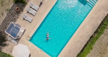 Top down view of a woman in a blue swimsuit lying on her back in the pool, slow motion. Relaxing concept, summer lifestyle. Camera descend and rotates counterclockwise