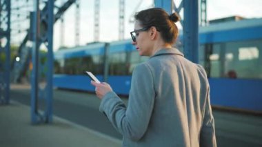 Sabahın erken saatlerinde şehirde dolaşan ve arka planda tramvay ve akıllı telefon kullanan beyaz iş kadını. Arkadan bak. İletişim, iş günü, yoğun yaşam konsepti.