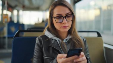 Public transport. Woman in glasses in tram using smartphone chatting and texting with friends, slow motion. City, urban, transportation.