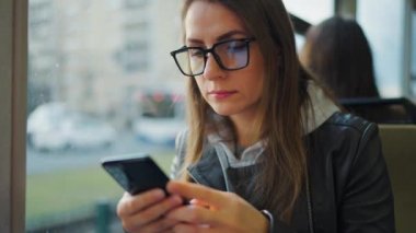 Public transport. Woman in glasses in tram using smartphone chatting and texting with friends, slow motion. City, urban, transportation.