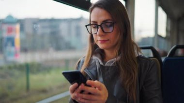 Public transport. Woman in glasses in tram using smartphone chatting and texting with friends, slow motion. City, urban, transportation.