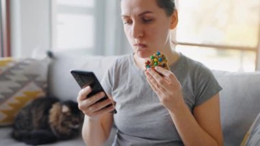 Renkli çipli kurabiye yiyip aynı anda akıllı telefon kullanan bir kadın. Sağlıksız yemek alışkanlıkları konsepti. Gıda kalorisi