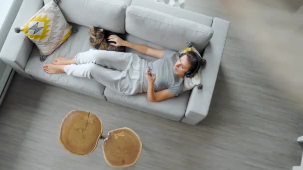 헤드폰을 위에서 소파에 누워서 스마트폰을 사용하고 고양이를 쓰다듬고 — 비디오