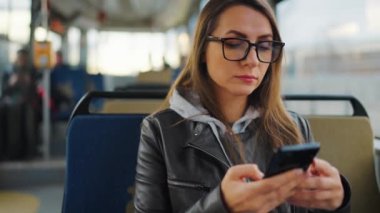 Public transport. Woman in glasses in tram using smartphone chatting and texting with friends, slow motion. City, urban, transportation.