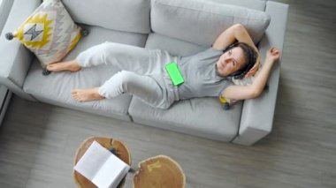 Overhead shot of relaxed woman listening to music in headphones on smartphone with green mock-up screen, lying on couch at home