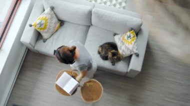 Kulaklıklı kadının yukarıdan çekilmiş fotoğrafı. Evdeki koltuğa uzanmış, akıllı telefon kullanıyor ve pofuduk bir kediyi okşuyor. 