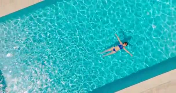 青い水着の女性のトップダウンビューは プール スローモーションで彼女の背中に横たわって カメラが立ち上がり 時計回りに回転します リラックスしたコンセプト 夏のライフスタイル — ストック動画