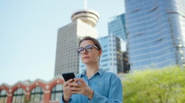 Gözlüklü beyaz kadın şehirde dolaşıyor ve akıllı telefon kullanıyor, ağır çekimde. Arka planda gökdelenler var. İletişim, iş günü, yoğun yaşam konsepti.