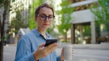 Gözlüklü beyaz kadın şehirde dolaşıyor, akıllı telefon kullanıyor ve kahve içiyor, ağır çekimde. Arka planda gökdelenler var. İletişim, iş günü, yoğun yaşam konsepti.