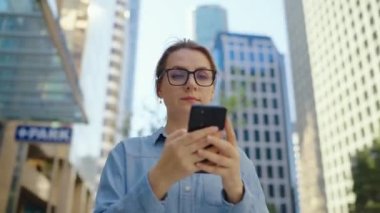Gözlüklü beyaz kadın şehirde dolaşıyor ve akıllı telefon kullanıyor. Arka planda gökdelenler var. İletişim, iş günü, yoğun yaşam konsepti.