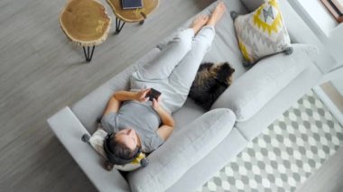 Kulaklıklı kadının yukarıdan çekilmiş fotoğrafı. Evdeki koltuğa uzanmış, akıllı telefon kullanıyor ve pofuduk bir kediyi okşuyor. 