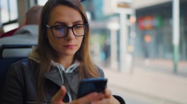 Public transport. Woman in glasses in tram using smartphone chatting and texting with friends, slow motion. City, urban, transportation.