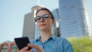 Gözlüklü beyaz kadın şehirde dolaşıyor ve akıllı telefon kullanıyor, ağır çekimde. Arka planda gökdelenler var. İletişim, iş günü, yoğun yaşam konsepti.