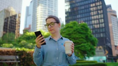 Gözlüklü beyaz kadın şehirde dolaşıyor, akıllı telefon kullanıyor ve kahve içiyor, ağır çekimde. Arka planda gökdelenler var. İletişim, iş günü, yoğun yaşam konsepti.