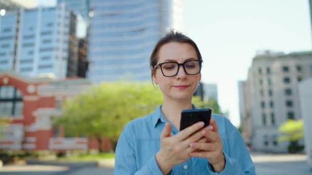 戴眼镜的白人女人在城市里走来走去 用智能手机 慢动作 摩天大楼在后面 工作日 忙碌生活概念 — 图库视频影像