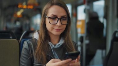 Public transport. Woman in glasses in tram using smartphone chatting and texting with friends, slow motion. City, urban, transportation.