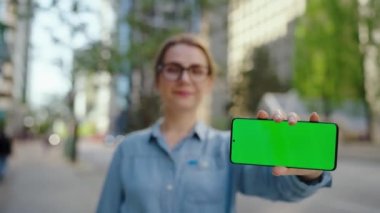 Gözlüklü kadın şehir caddesinde duruyor ve kameraya yeşil model ekranlı akıllı telefonu gösteriyor. Arka planda gökdelenler var. İletişim, iş günü, yoğun yaşam konsepti.