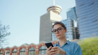 Gözlüklü beyaz kadın şehirde dolaşıyor ve akıllı telefon kullanıyor, ağır çekimde. Arka planda gökdelenler var. İletişim, iş günü, yoğun yaşam konsepti.