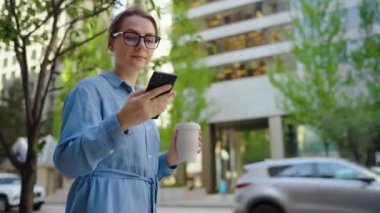 Gözlüklü beyaz kadın şehirde dolaşıyor, akıllı telefon kullanıyor ve kahve içiyor, ağır çekimde. Arka planda gökdelenler var. İletişim, iş günü, yoğun yaşam konsepti.