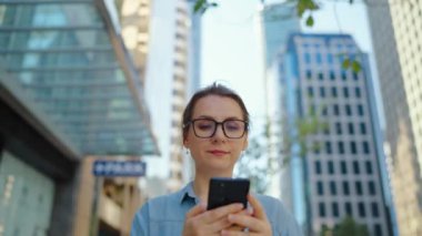 Gözlüklü beyaz kadın şehirde dolaşıyor ve akıllı telefon kullanıyor, ağır çekimde. Arka planda gökdelenler var. İletişim, iş günü, yoğun yaşam konsepti.