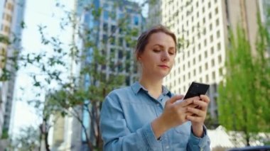 Gözlüklü beyaz kadın şehirde dolaşıyor ve akıllı telefon kullanıyor, ağır çekimde. Arka planda gökdelenler var. İletişim, iş günü, yoğun yaşam konsepti.