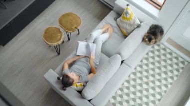 Overhead view of woman reading the book, lying on couch at home. Fluffy cat sitting next to her