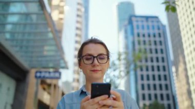 Gözlüklü beyaz kadın şehirde dolaşıyor ve akıllı telefon kullanıyor. Arka planda gökdelenler var. İletişim, iş günü, yoğun yaşam konsepti.