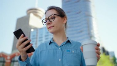 Gözlüklü beyaz kadın şehirde dolaşıyor, akıllı telefon kullanıyor ve kahve içiyor, ağır çekimde. Arka planda gökdelenler var. İletişim, iş günü, yoğun yaşam konsepti.