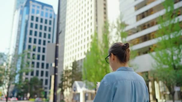街中を歩いているメガネ姿の白人女性やスマートフォン バックビューを使用しています スローモーション 高層ビルが背景にあります コミュニケーション 仕事の日 忙しい生活の概念 — ストック動画