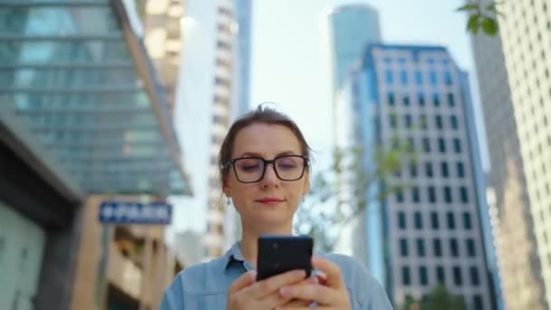 안경을 도시를 돌아다니며 스마트폰을 사용하고 있습니다 마천루들 배경에 있습니다 의사소통 — 비디오