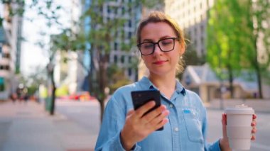 Gözlüklü beyaz kadın sokakta duruyor, akıllı telefon kullanıyor ve kahve içiyor. Arka planda gökdelenler var. İletişim, iş günü, yoğun yaşam konsepti.