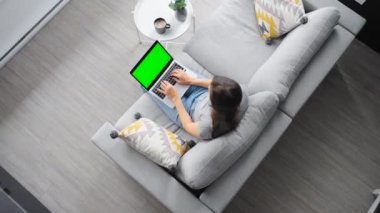 Yeşil maket ekranlı dizüstü bilgisayar kullanan bir kadının evdeki kanepede otururken çekilen resmi..
