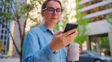 Gözlüklü beyaz kadın şehirde dolaşıyor, akıllı telefon kullanıyor ve kahve içiyor, ağır çekimde. Arka planda gökdelenler var. İletişim, iş günü, yoğun yaşam konsepti.