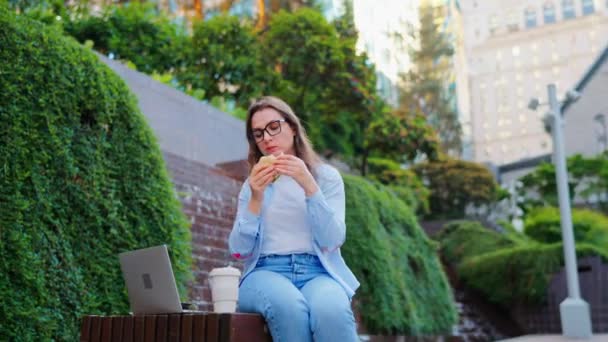 サンドイッチとコーヒーでランチをしながら ラップトップを持って公園に座っている白人女性 アウトドア 女性のフリーランスは彼女のラップトップと一緒に働きます — ストック動画