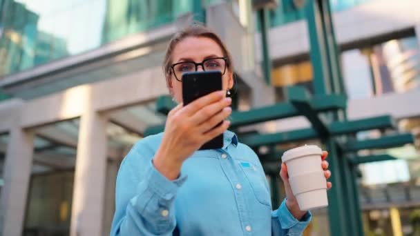 안경을 여성은 스마트폰과 커피를 사용하여 거리에 있습니다 마천루는 배경에 있습니다 — 비디오