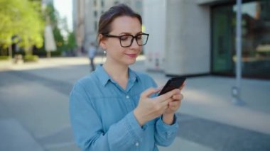 Gözlüklü beyaz kadın şehirde dolaşıyor ve akıllı telefon kullanıyor. Arka planda gökdelenler var. İletişim, iş günü, yoğun yaşam konsepti.