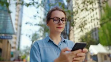 Gözlüklü beyaz kadın şehirde dolaşıyor ve akıllı telefon kullanıyor. Arka planda gökdelenler var. İletişim, iş günü, yoğun yaşam konsepti.