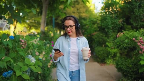 Gözlüklü Kulaklıklı Elinde Kahve Akıllı Telefonuyla Yaz Günü Yeşil Bir — Stok video