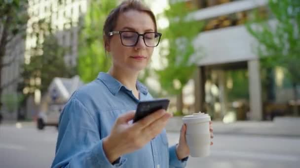 안경을 스마트폰을 사용하고 커피를 마시면서 도시를 걸어다니고 있습니다 마천루들 배경에 — 비디오
